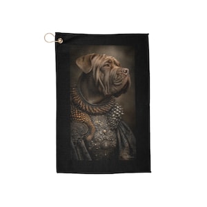 Puede incluir: Toalla de golf negra con un retrato de un mastín napolitano. El perro lleva una armadura decorativa y un collar de púas. La toalla tiene un ojal dorado para colgar. Tema real.