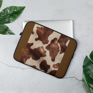 May include: A laptop sleeve featuring a cowhide pattern in shades of brown and cream. The sleeve has a brown border and a small heart-shaped embellishment. The laptop sleeve is designed to protect a laptop.
