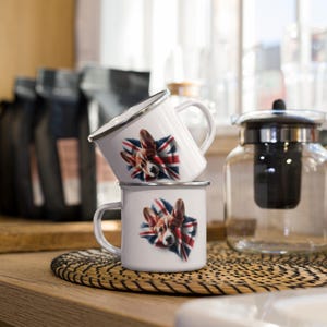 Op de afbeelding: Twee witte emaille mokken op elkaar gestapeld. Elke mok heeft een afbeelding van een Corgi-hond met de Union Jack-vlag. De mokken hebben een zilveren rand en een wit handvat. De mokken staan op een geweven mat.