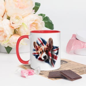 Op de afbeelding: Witte keramische mok met een rode binnenkant en handvat. De mok heeft een afbeelding van een Corgi-hond met de Union Jack-vlag op de achtergrond. De mok wordt omringd door bloemen, snoep en chocolade.