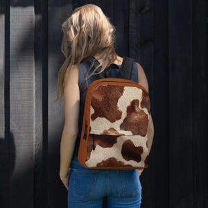 May include: A brown and white cowhide-patterned backpack with a front pocket and black straps. The backpack has a brown border and a black zipper. The person in the image is wearing blue jeans and a gray top.