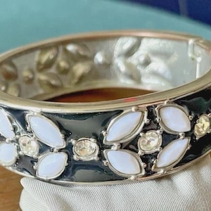 May include: A hinged bangle bracelet featuring a polished silver-tone metal frame with a glossy black enamel inlay. The surface is decorated with repeating floral motifs composed of white marquise-shaped cabochons and clear round rhinestones.