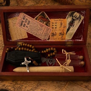 May include: A rectangular wooden box with a red interior containing a black coffin-shaped box, a wooden stake, two glass vials with cork stoppers, a rosary with brown beads, and a silver crucifix. The lid features paper inserts with text.
