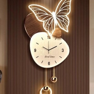 May include: A wall clock featuring an irregular cream-colored face with black numerals, the text "Good times," and black hands. Above the face is a glowing white butterfly outline. Below, two gold-toned pendulums hang, one with a ring and one with a sphere.