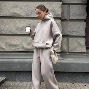 Può includere: Un completo composto da felpa con cappuccio grigio chiaro e pantaloni della tuta a gamba larga abbinati. La felpa con cappuccio ha una tasca frontale e un cappuccio. I pantaloni sono ampi. L'outfit è abbinato a scarpe da ginnastica bianche e una piccola borsa beige. La persona tiene in mano una tazza di caffè.