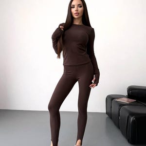 Puede incluir: Conjunto marrón de top de manga larga y leggings a juego. El top tiene cuello redondo y mangas ajustadas. Los leggings tienen un diseño de estribo en los tobillos. El conjunto está hecho de un material suave y elástico. La persona lleva zapatos de ante grises.
