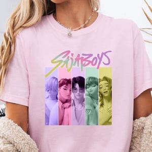 Puede incluir: Una camiseta rosa claro con el texto "SajaBoys" en una fuente colorida y estilizada. Debajo del texto hay seis im&aacute;genes de personas, cada una sobre una franja vertical de diferente color. La camiseta est&aacute; hecha de un material suave.