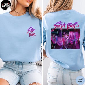 May include: Light blue sweatshirt with the text "Saja Boys" in a gradient pink and purple font on the front and back. The back features a graphic of four stylized faces in a rectangular frame.