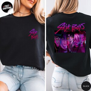 May include: Black sweatshirt with the text "Saja Boys" in a gradient purple and pink font on the front and back. The back features a graphic of four figures in a rectangular frame, also in purple and pink.