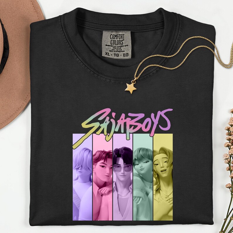 Puede incluir: Camiseta negra con el texto "SajaBoys" en una fuente colorida y estilizada. La camiseta presenta un gr&aacute;fico de cinco figuras de dibujos animados en paneles verticales de colores. Un collar con colgante de estrella dorada descansa sobre la camiseta.