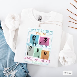 May include: White sweatshirt with a graphic design featuring four pop art portraits and the text "I WAS THERE THEN MILLENNIUM AND I'M HERE NOW". A silver necklace with a pendant is draped on the sweatshirt. The sweatshirt is folded on a white surface.