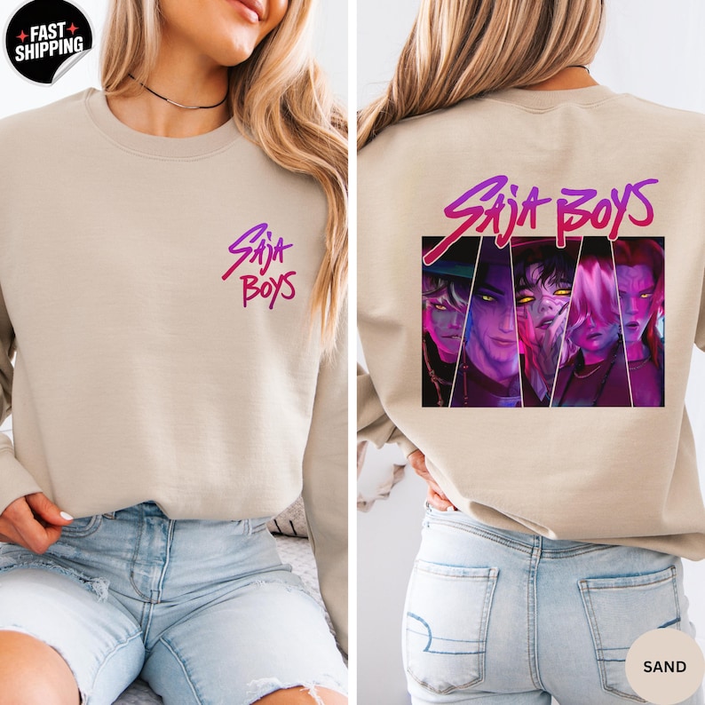 May include: Sand-colored sweatshirt with the text "Saja Boys" in a gradient purple and pink font on the front and back. The back features a graphic of four stylized faces. The sweatshirt is made of a soft material.