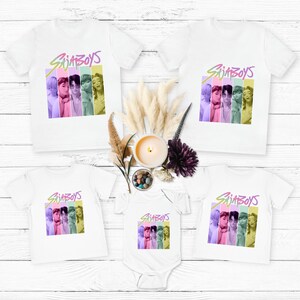Puede incluir: Camisetas blancas y un body con un gr&aacute;fico colorido de un grupo de personas con el texto "SajaBoys". El gr&aacute;fico est&aacute; en un dise&ntilde;o de rayas verticales con colores pastel. Las camisas son de manga corta.
