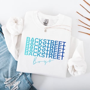 May include: White sweatshirt with the text "BACKSTREET" stacked in a blue gradient, with "Boys" in a script font below. A silver necklace with a small pendant is on the sweatshirt. The sweatshirt is on a white surface with a pair of blue jeans.
