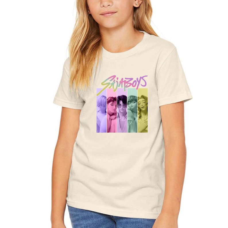 Puede incluir: Camiseta color crema con el texto "SAJA BOYS" en una fuente colorida y estilizada. Debajo, seis franjas verticales, cada una con un color diferente y la imagen de una persona. La camiseta es de material suave.