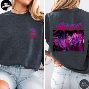 May include: Dark heather gray sweatshirt with the text "Sia Boys" in a pink and purple gradient on the front and back. The back features a graphic of four stylized faces in a rectangular frame.