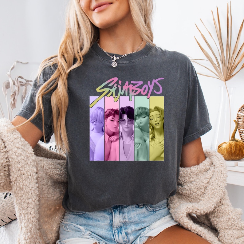 Puede incluir: Camiseta gris oscuro con el texto "Saja Boys" en degradado arco&iacute;ris sobre un gr&aacute;fico de seis personas en bloques de color verticales. La camiseta es de estilo casual.