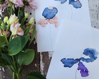 May include: Two watercolor paintings of abstract flowers in shades of blue, purple, and pink on white paper.