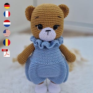 Op de afbeelding: Een handgemaakte gehaakte teddybeer met een lichtbruin lichaam, gekleed in een blauwe jumpsuit met een gerimpelde kraag. De beer heeft zwarte ogen, een witte snuit en witte schoenen. De afbeelding bevat vlaggen en een PDF-pictogram.