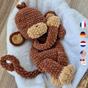 May include: A handmade brown crochet monkey toy with a tan face, ears, and feet, lying on a white fluffy blanket. The monkey has closed eyes, suggesting it is sleeping. The tail is curled. A soft, cuddly plush toy.
