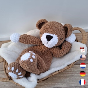 May include: A handmade brown crochet teddy bear with white accents on its paws and muzzle, resting on a fluffy white blanket inside a woven basket. The bear has closed eyes, suggesting sleep.