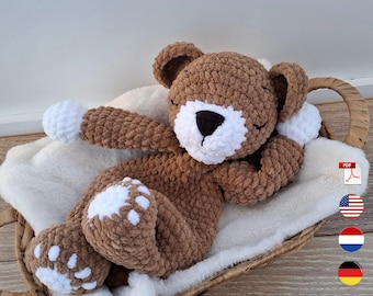 May include: A handmade brown crochet teddy bear with white accents on its paws and muzzle, resting on a fluffy white blanket inside a woven basket. The bear has closed eyes, suggesting sleep.