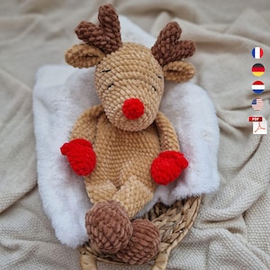 May include: A crocheted reindeer plush toy made of tan yarn with a textured finish. It features dark brown antlers, a bright red nose, and red mittens on its hands. The reindeer has closed eyes stitched in black thread and round brown feet.