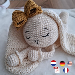 May include: A cream-colored crocheted bunny toy with a mustard yellow bow. The bunny has closed eyes and a sweet expression. The toy is handmade and appears soft and cuddly, perfect for a baby or child.