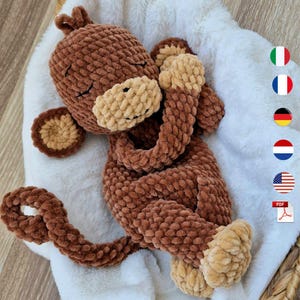 May include: A brown and beige crocheted monkey plush toy with a soft, textured yarn finish. The monkey has closed eyes, a rounded snout, and a long, curled tail. It is crafted from plush yarn, featuring a dark brown body with lighter beige accents on the face, ears, hands, and feet, presented in a sleeping pose.