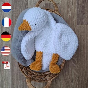 May include: A handmade white crochet goose with an orange beak and feet, resting in a woven basket. The plush toy has closed eyes and is set against a gray, fluffy background.