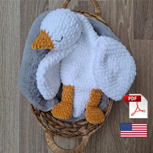 May include: A handmade white crochet goose plush toy with orange beak and feet, resting in a woven basket with a grey faux fur blanket. The goose has closed eyes. A PDF icon and the American flag are in the corner.