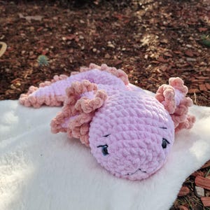 Könnte beinhalten: Ein gehäkeltes Axolotl-Stofftier aus weichem, hellrosa Garn. Das Wesen hat einen abgerundeten Kopf, dunkle gestickte Augen und gerüschte, pfirsichfarbene Kiemen, die sich an den Kopfseiten und entlang des langen, segmentierten Körpers erstrecken.