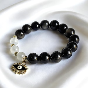 Può includere: Un braccialetto di perline con perline di pietre preziose nere e bianche. Il braccialetto include un ciondolo color oro con un disegno a occhio. Le perline sono infilate su un elastico.