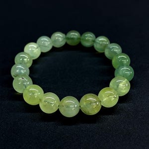 Könnte beinhalten: Ein Armband aus runden, hellgrünen Prehnit-Perlen. Die Perlen haben eine durchscheinende Qualität und variieren in Grüntönen. Das Armband wird vor einem dunklen Hintergrund präsentiert, um die Farbe und Textur des Edelsteins hervorzuheben.