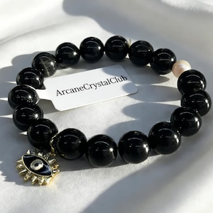 Può includere: Un braccialetto di perline nere con un ciondolo color oro a forma di occhio. Il braccialetto include un'etichetta con la scritta "ArcaneCrystalClub". Le perline sono rotonde e lucide, con alcune perline di colore più chiaro.