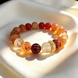 Może przedstawiać: Bransoletka z kolorowych kamieni szlachetnych. Zawiera pomarańczowe, czerwone i przezroczyste koraliki oraz złoty element dekoracyjny. Bransoletka jest prezentowana na białej, jedwabistej tkaninie.
