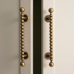 May include: A pair of antique brass door handles. Each handle has a string of small, round beads leading to a larger ball at each end. They are mounted on a white door, with a grey door visible on the left.