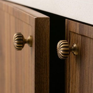 Może przedstawiać: Zbliżenie dwóch drewnianych drzwi szafki z mosiężnymi, żebrowanymi gałkami. Drzwi są w ciepłym brązowym kolorze, a gałki mają wygląd vintage. Biały blat jest widoczny nad szafkami.