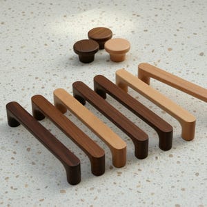 Könnte beinhalten: Eine Auswahl an Holzmöbelbeschlägen, darunter Knöpfe und Griffe. Die Knöpfe sind rund, während die Griffe rechteckig mit gebogenen Griffen sind. Die Holztöne variieren von hell- bis dunkelbraun und zeigen natürliche Maserungsmuster. Das Hardware ist für Möbel.