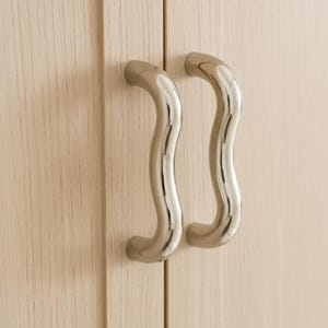Könnte beinhalten: Nahaufnahme von zwei polierten, gebogenen Metallschrankgriffen. Die Griffe haben eine wellenförmige, längliche S-Form und sind an einer hellen Holzschranktür montiert. Die Griffe sind silbern glänzend.
