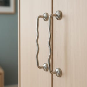 Könnte beinhalten: Nahaufnahme von zwei Schrankgriffen aus gebürstetem Nickel. Die Griffe haben ein wellenförmiges, organisches Design und sind an einem hellen Holzschrank montiert. Die Griffe sind ein modernes, minimalistisches Design.