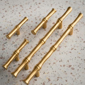 Może przedstawiać: Złote uchwyty do szafek w stylu bambusowym. Uchwyty mają teksturowany, segmentowy design, przypominający łodygi bambusa. Okucia są ułożone na nakrapianej, neutralnej powierzchni. Uchwyty różnią się rozmiarem.