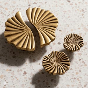 Könnte beinhalten: Satz von vier messingfarbenen Schrankknöpfen und -griffen. Zwei größere Griffe sind fächerförmig, und zwei kleinere Knöpfe sind kreisförmig mit radialem Design. Die Hardware hat eine Vintage-Ästhetik mit warmem, metallischem Finish. Die Knöpfe und Griffe werden auf einer gesprenkelten Oberfläche präsentiert.