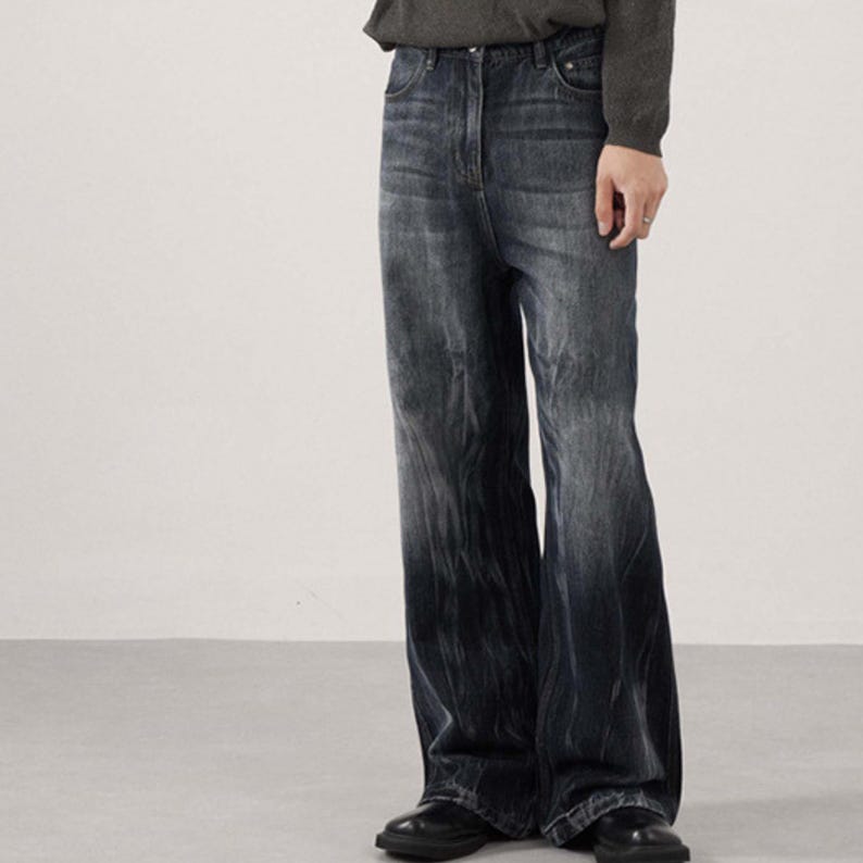 May include: Dark wash denim jeans with a wide leg design. The jeans have a faded effect, transitioning from a lighter shade at the top to a darker shade towards the bottom. The jeans are paired with black shoes.