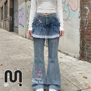 Puede incluir: Vaqueros de mezclilla azul claro con diseño de pernera acampanada. Los vaqueros presentan una superposición de falda de mezclilla con ribete de encaje y bordado floral. Un gráfico de ratón de dibujos animados y texto están impresos en la parte delantera de los vaqueros. La modelo lleva una camisa blanca de manga larga.