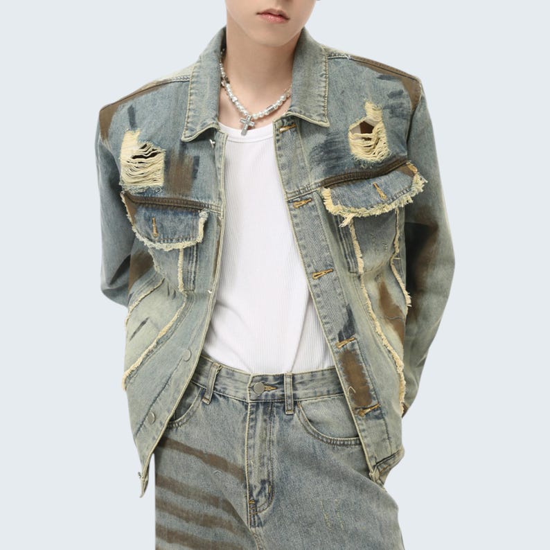 May include: Distressed denim jacket with ripped details and brown patches. The jacket has two chest pockets and a button-down front. The model is wearing a white undershirt and matching jeans.