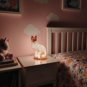 Könnte beinhalten: Eine katzenförmige Lampe, die warmes Licht ausstrahlt, steht auf einem weißen Nachttisch. Die Lampe hat ein Muster. Das Kinderzimmer hat rosa Wände, Wolkenaufkleber und gerahmte Bilder, was eine gemütliche Atmosphäre schafft.
