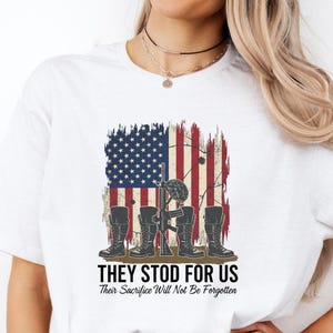 May include: White t-shirt featuring a patriotic design with an American flag and military boots. The text "THEY STOD FOR US" and "Their Sacrifice Will Not Be Forgotten" is printed below the graphic. A casual, everyday wear tee.