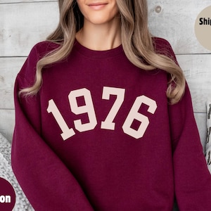 May include: Maroon sweatshirt with the year "1976" printed in a cream-colored font. The sweatshirt is a solid color and has a crew neck. The text "Maroon" is in a circle in the lower left corner.
