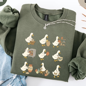 May include: Military green sweatshirt featuring a whimsical illustration of ducks engaged in pottery. The design includes ducks working at a pottery wheel, carrying pots, and arranging finished pieces. A silver necklace is draped across the sweatshirt.